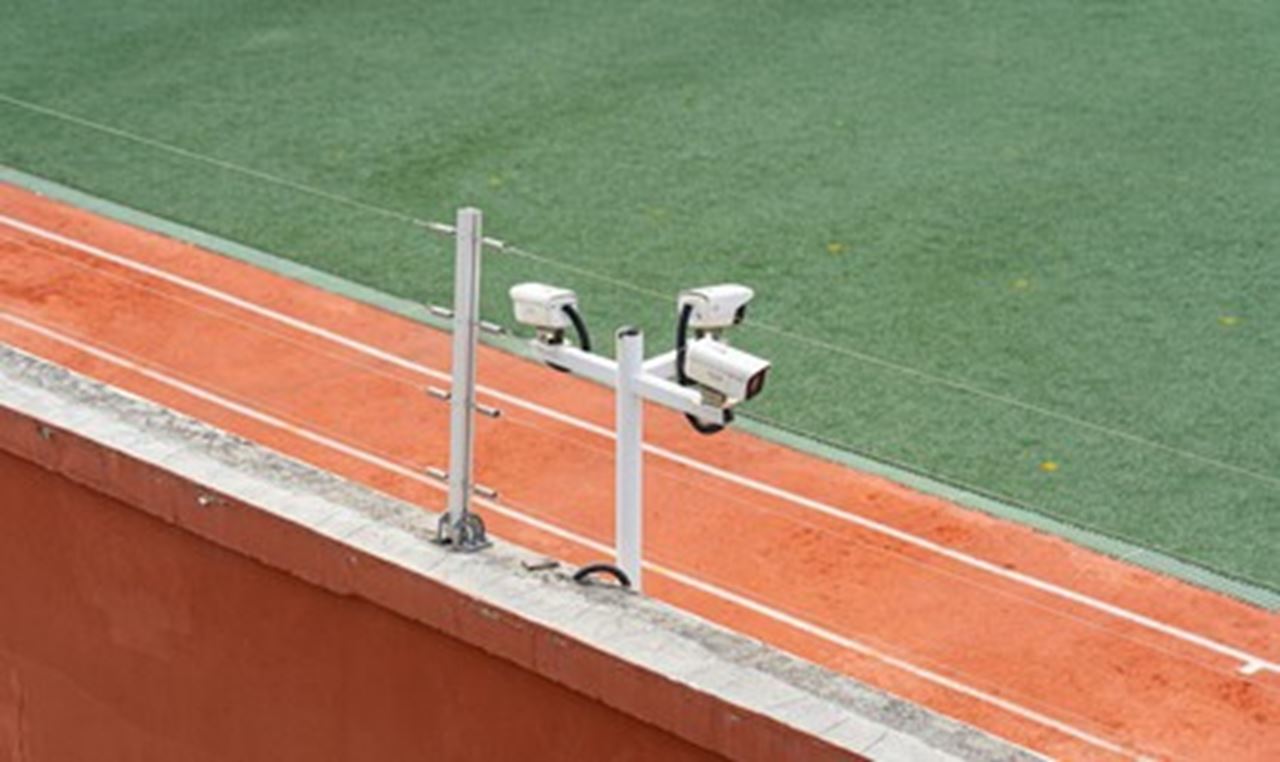Deux caméras de surveillance fixées sur un poteau blanc, situées en bordure d'un terrain de sport avec une piste rouge et du gazon synthétique vert en arrière-plan.