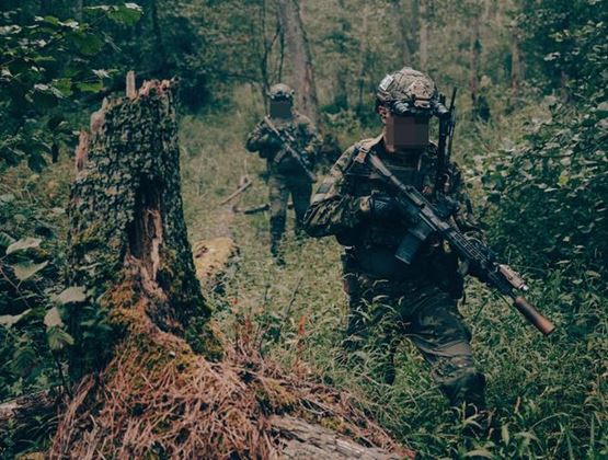 Two soldiers equipped with tactical gear advancing through a dense forest during a training exercise