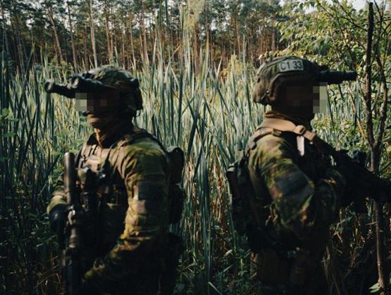 Two soldiers in camouflage gear stand back to back in a dense, green forest area, equipped with rifles and night vision goggles.