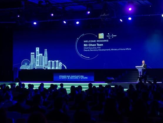 Conference stage at a technology exhibition, with a speaker at a podium and a screen showing welcome remarks, blue lighting and audience in the foreground.