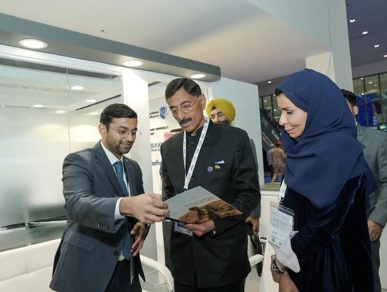 Exchange between representatives at a trade show: an exhibitor presenting a brochure to two official visitors at a modern booth, all wearing accreditation badges.
