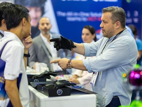 Sur un stand de salon professionnel, un exposant portant un gant noir présente et explique un dispositif à un visiteur.