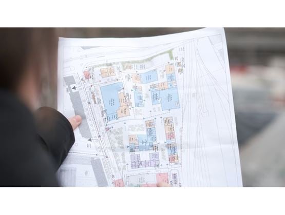 A person holds a detailed, color-coded architectural site plan showing the layout of various buildings and areas, likely outdoors at a construction site.