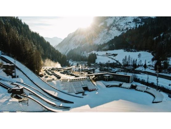 Aerial view of a ski jumping hill with snow-covered spectator stands and nearby buildings, set in a mountain valley during winter, with the sun rising behind the peaks
