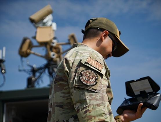 Militaire en uniforme tenant une télécommande, devant un équipement de surveillance ou de défense, sous un ciel bleu.
