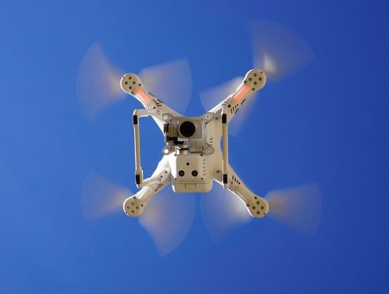 Drone blanc en vol vu de dessous, avec hélices en mouvement et caméra embarquée, sur fond de ciel bleu.