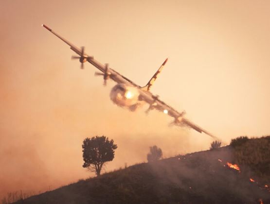 Avion de lutte contre les incendies volant à basse altitude au-dessus d’un feu de forêt, avec flammes et fumée sur la colline.
