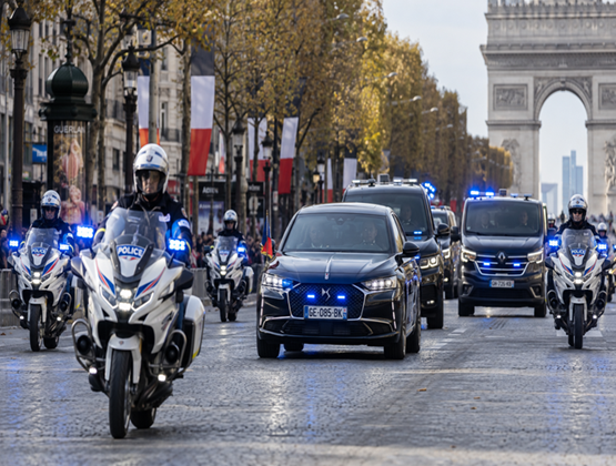 Police motorcycles escorting a motorcade along the Champs-Élysées, with blue lights flashing and the Arc de Triomphe behind them.