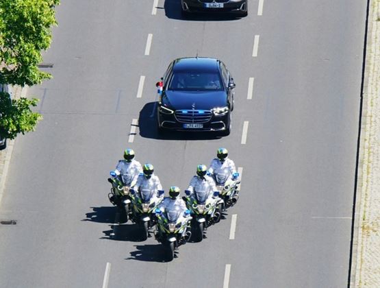Vue en plongée de cinq motos de police roulant en formation devant une voiture noire au sein d’un cortège.