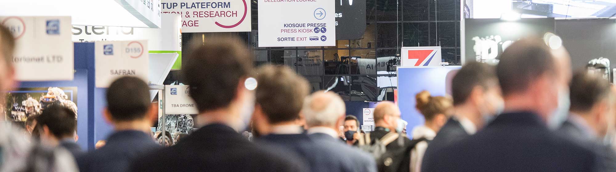 Milipol's trade show floor with various booths and a crowd of attendees. Some of the booths have banners with names like AVNON, also banners of the event such as :  Startup area or Innovation STAGE. The photo shows a busy and professional atmosphere.