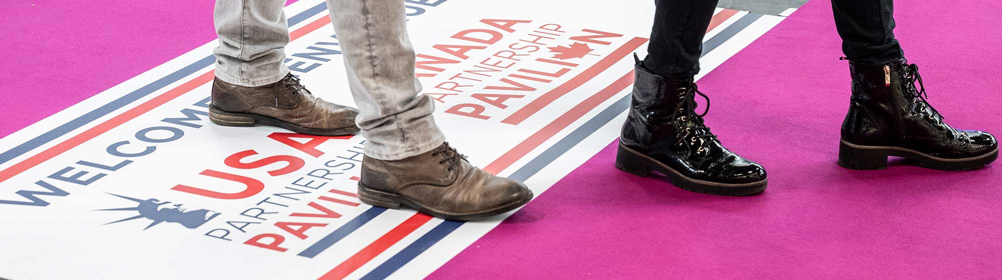 Zoom sur les jambes de deux visiteurs machant dans les allées, avec signalétique au sol indiquant le pavillon USA