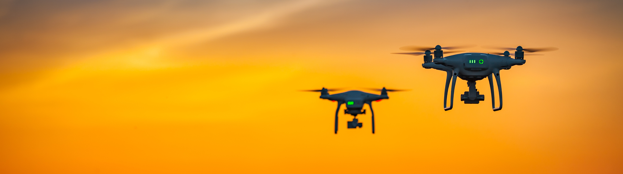Deux drones volant au coucher du soleil, en silhouette contre le ciel orange, symbolisant la surveillance aérienne.