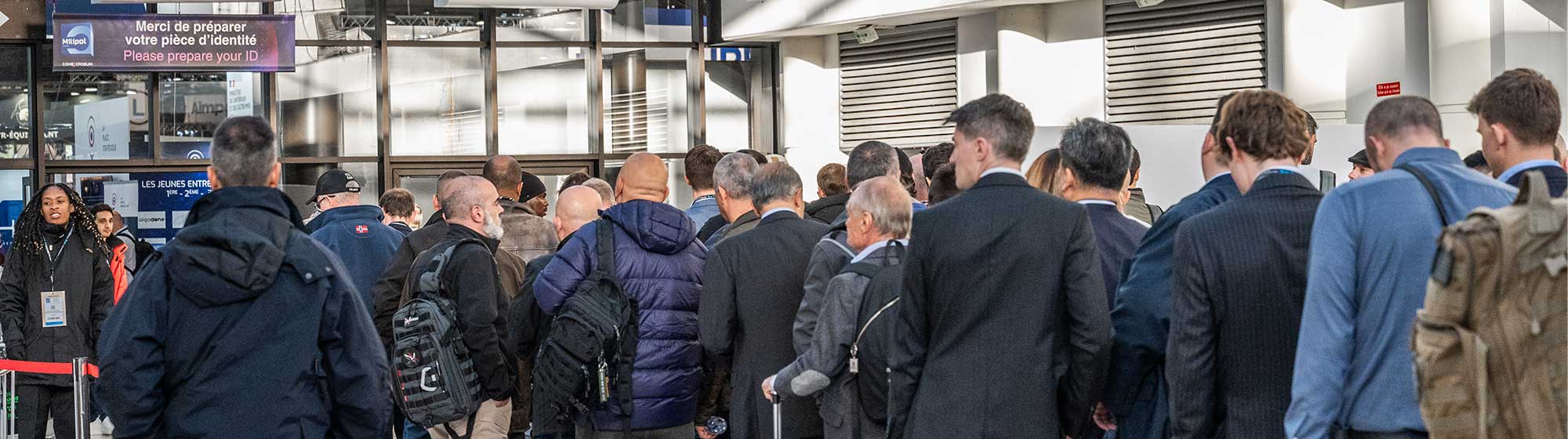 A group of people waiting in line at Milipol Paris's entrance under a sign that reads “Bienvenue Welcome” and instructs attendees to present their badges.  Attendees are lined up, waiting to enter.