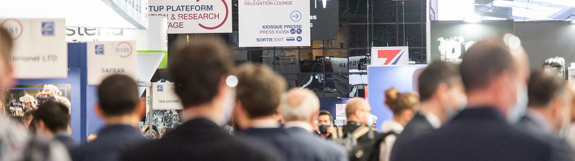 Salon Milipol avec divers stands et une foule de participants. Certains stands portent des bannières avec des noms comme AVNON, bannières de l'événement également affichées:  Zone startup, Innovation STAGE. La photo montre une atmosphère animée et professionnelle.