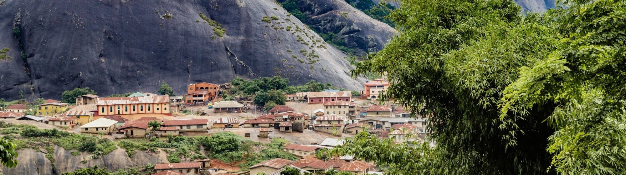 Vue panoramique d’un village nigérian entouré de montagnes rocheuses couvertes de végétation, sous un ciel nuageux.