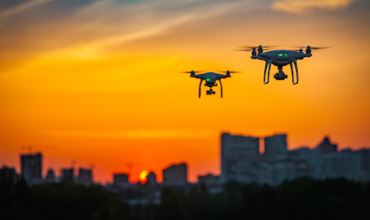 Deux drones volant au-dessus d’une ville au coucher du soleil, en silhouette contre le ciel orange, symbolisant la surveillance aérienne.