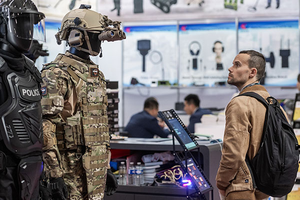 A man wearing a brown jacket and carrying a rucksack is observing two outfits on display, one a military fatigues, the other a police uniform with protection