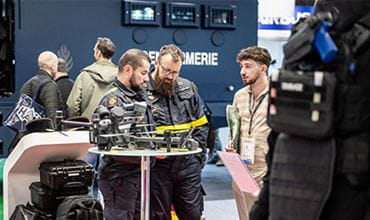 Deux hommes de la sécurité civile en uniformes militaires se tiennent autour d'une table auprès d’un visiteur 