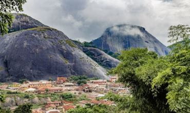 Vue panoramique d’un village nigérian entouré de montagnes rocheuses couvertes de végétation, sous un ciel nuageux.