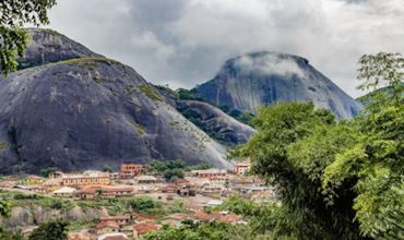Panoramic view of a Nigerian village surrounded by rocky mountains with greenery, under a cloudy sky.