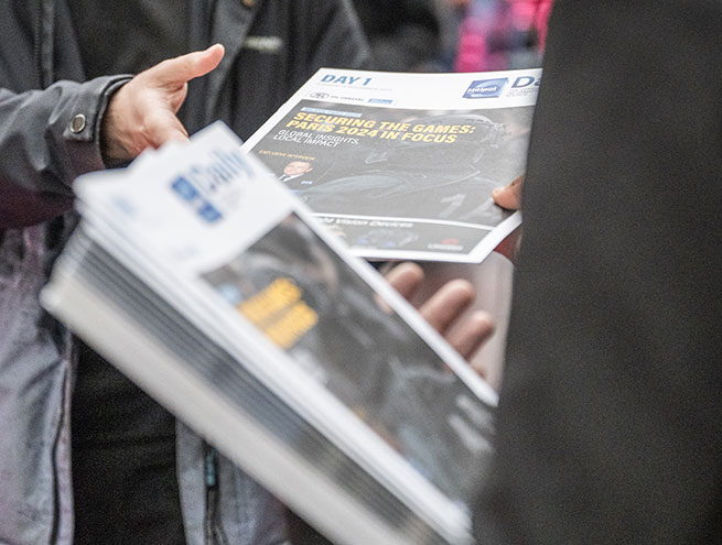 Une personne distribuant le daily news, le journal de Milipol Paris,  à une autre personne à l'entrée du salon