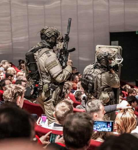 Simulated GIGN intervention in a crowded auditorium during Milipol Paris, with two armed tactical officers standing among the audience