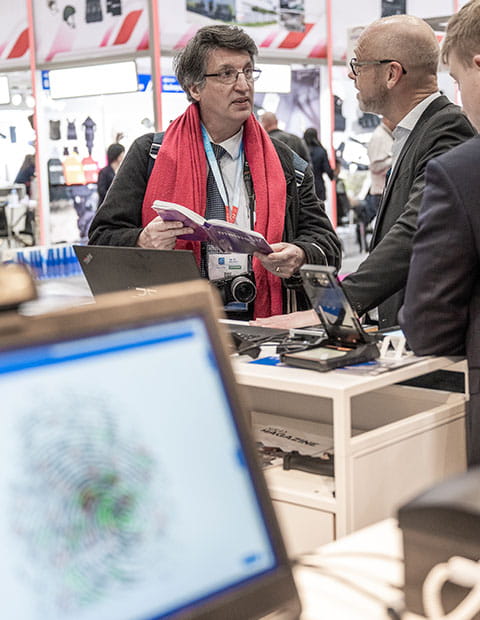 A visitor and an exhibitor chat together on a stand. A computer screen projects a digital imprint