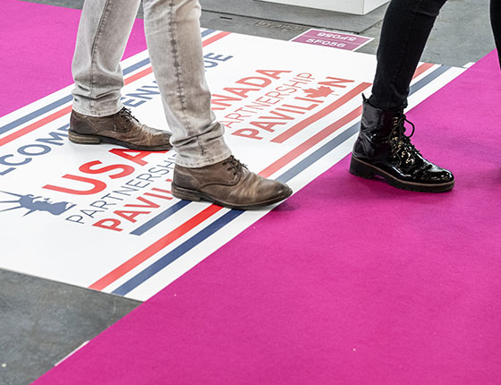 Zoom on the legs of two visitors walking through the aisles, with signs on the ground indicating the USA pavilion