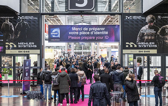 Crowded visitors' entrance hall with a Milipol welcome poster and two large posters advertising MEHLER SYSTEMS, a company that provides solutions for ballistic protection.