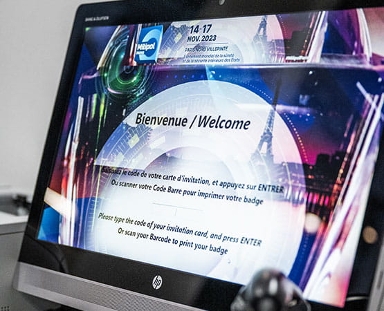 A computer monitor displaying a Milipol event welcome screen in both French and English, with instructions for printing a badge, placed on a desk with a keyboard and joystick.
