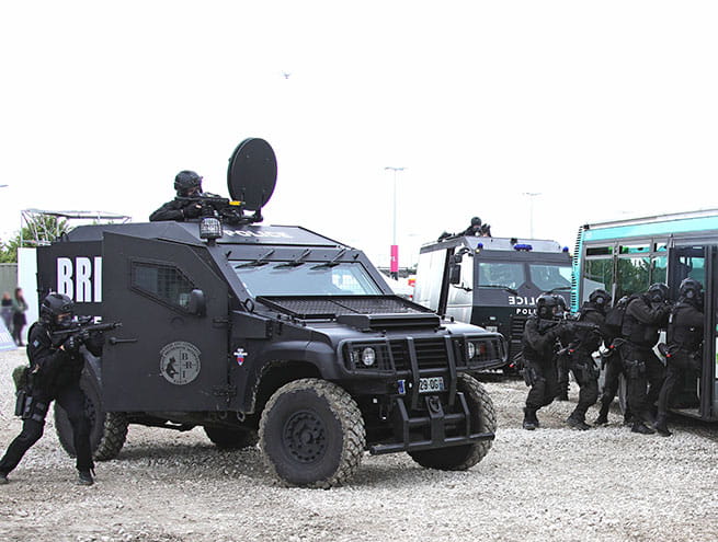 A group of armed police officers from the intervention brigade (BI/DOPC) in black uniforms stand next to a BRI vehicle and break into a bus.