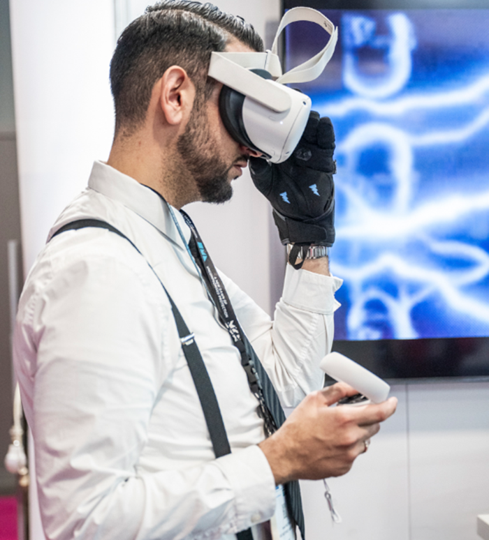 Homme en chemise blanche testant un casque de réalité virtuelle et des gants connectés sur un stand d’exposition.